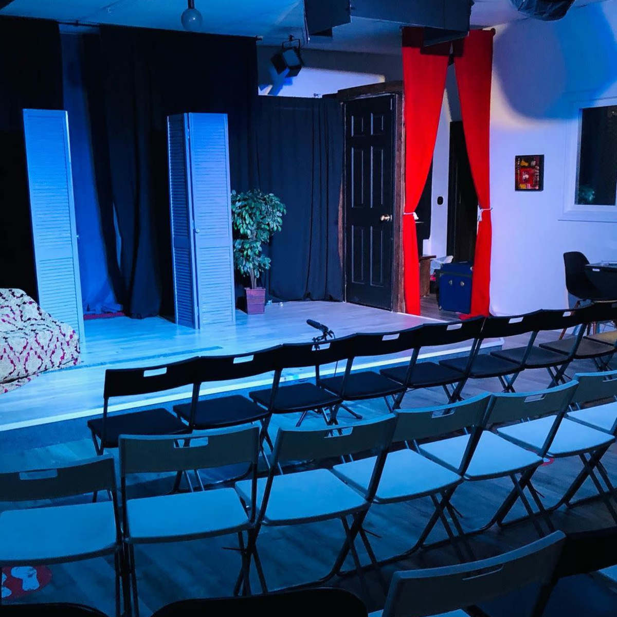 A theater stage with two folding screens, a plant in a pot, red curtains, and rows of empty black chairs arranged for an audience.