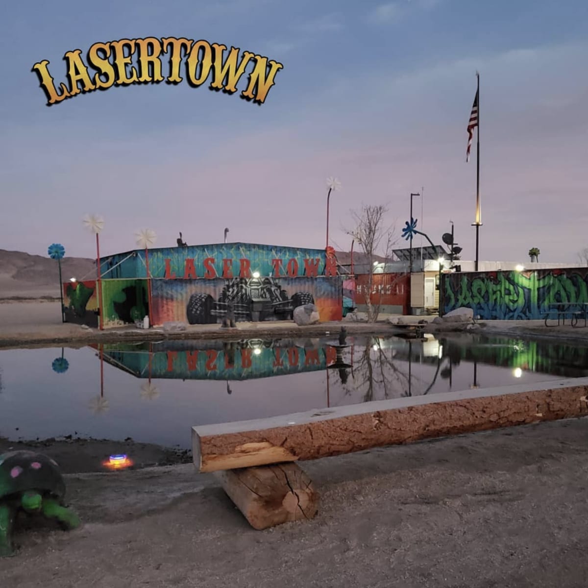 A colorful building labeled "LASERTOWN" near a small pond, with a flagpole and palm trees in the background.