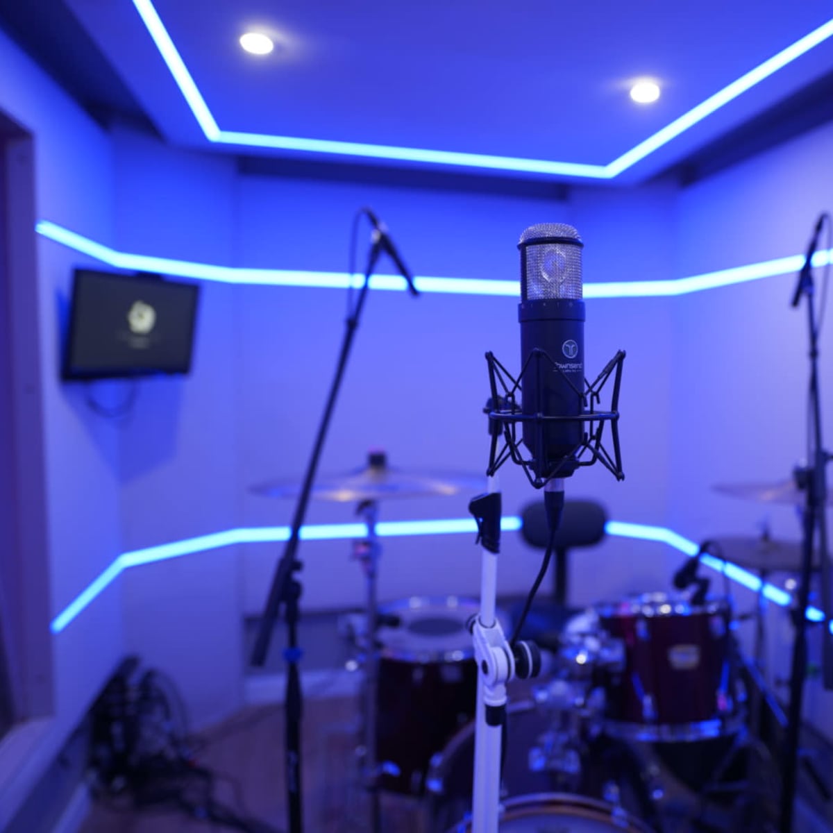 A microphone stands in a dimly lit recording studio with blue LED lights and a drum set in the background.