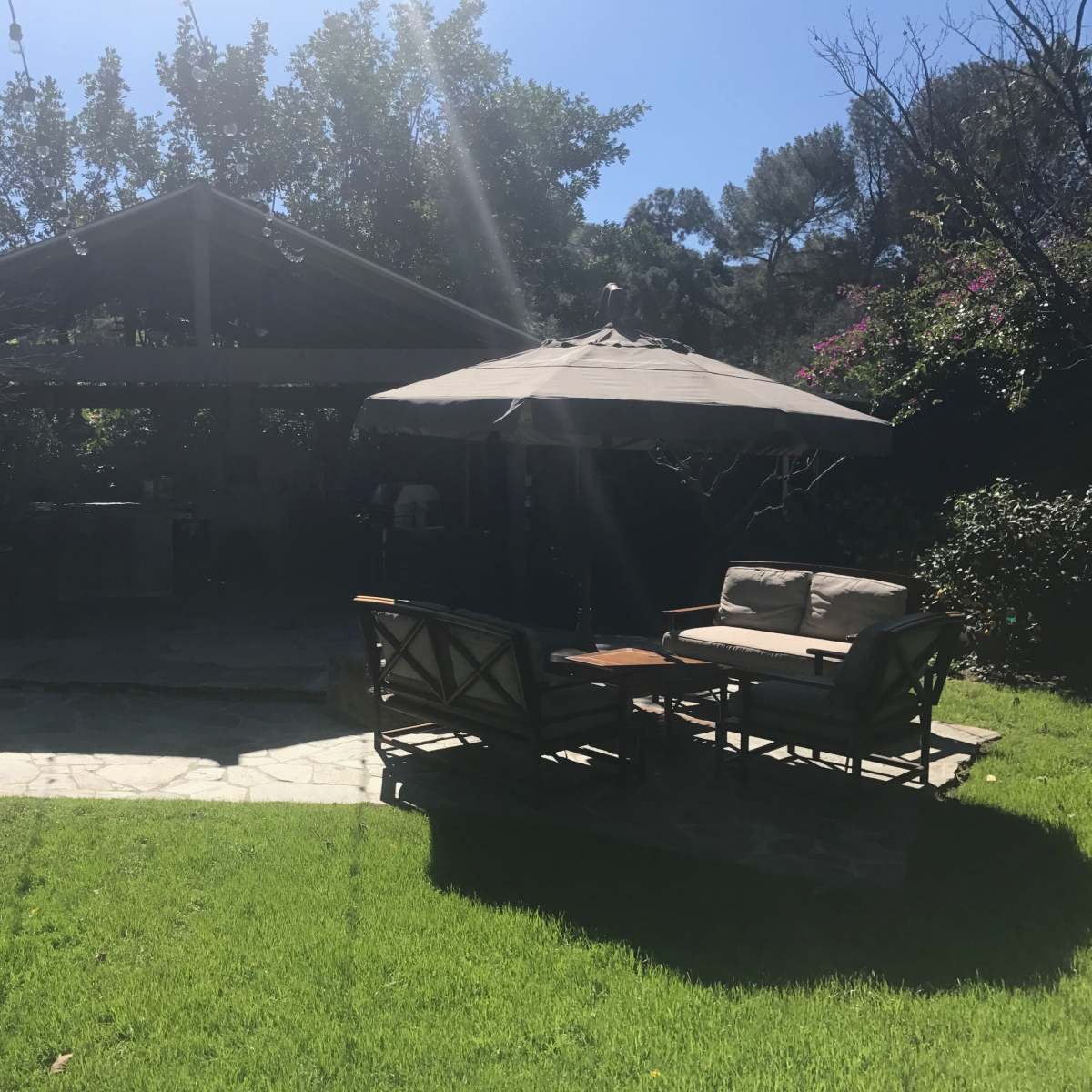 A shaded outdoor seating area with a sofa and table sits on a grassy lawn beside a stone pathway, leading to a covered patio surrounded by trees and flowering plants.