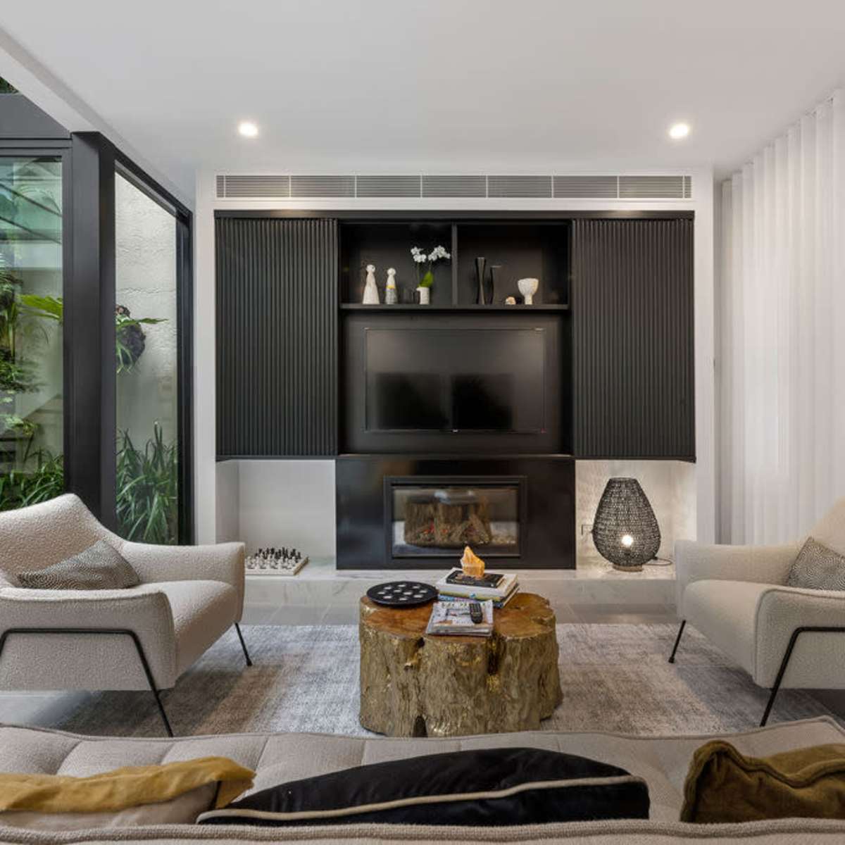 A modern living room with two armchairs, a wooden coffee table, a mounted television, and large windows allowing natural light.
