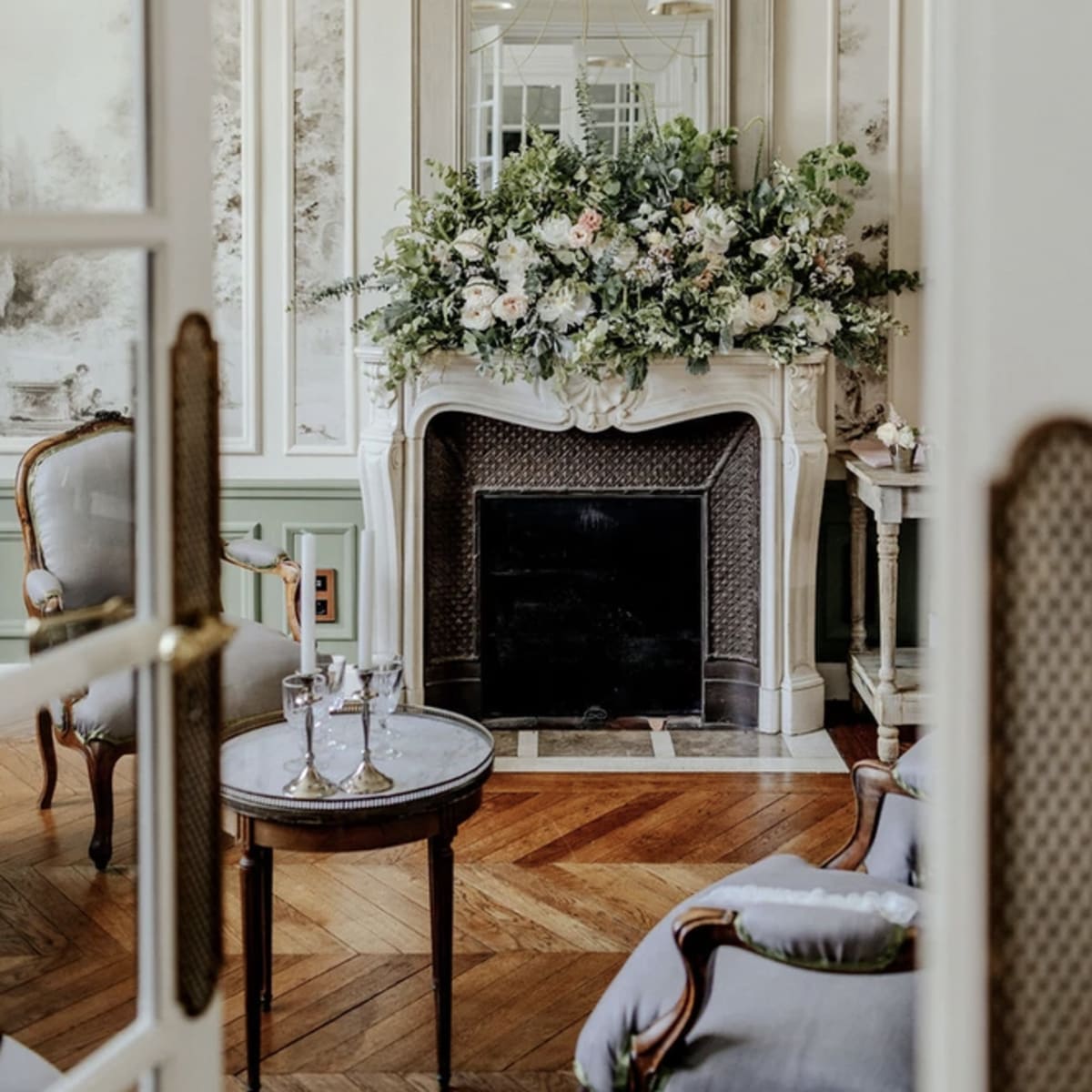 A well-decorated living room features a fireplace adorned with a floral arrangement, elegant furnishings, and a stylish chandelier.