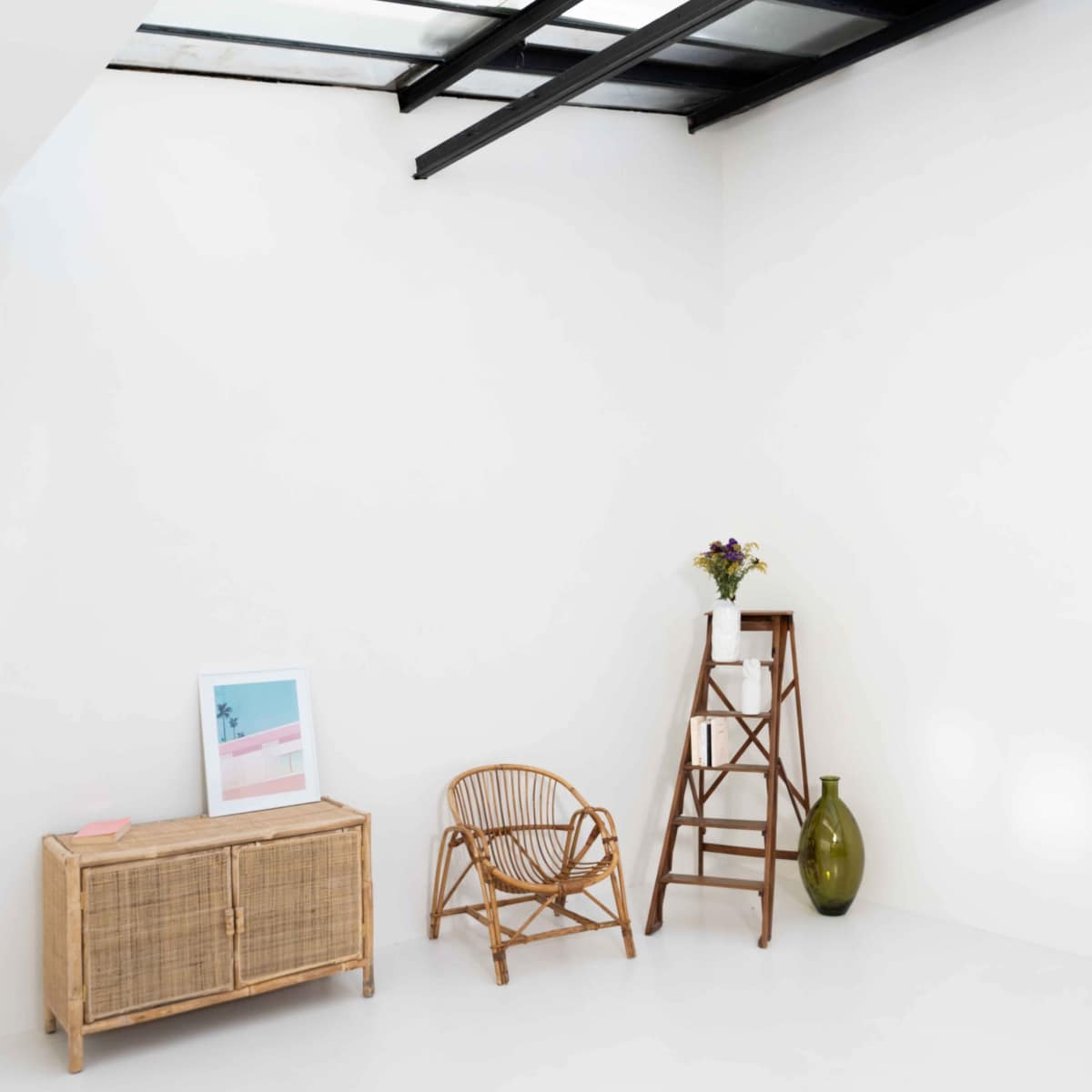 A minimalist interior space featuring a wicker chair, a wooden side cabinet, a ladder-style shelf with a vase, and a framed picture against a white wall.