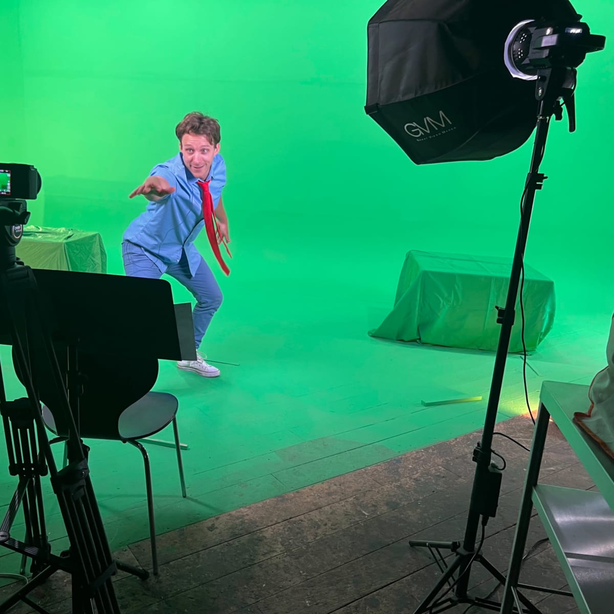 A person in a blue outfit and red tie poses playfully in front of a green screen, surrounded by filming equipment.