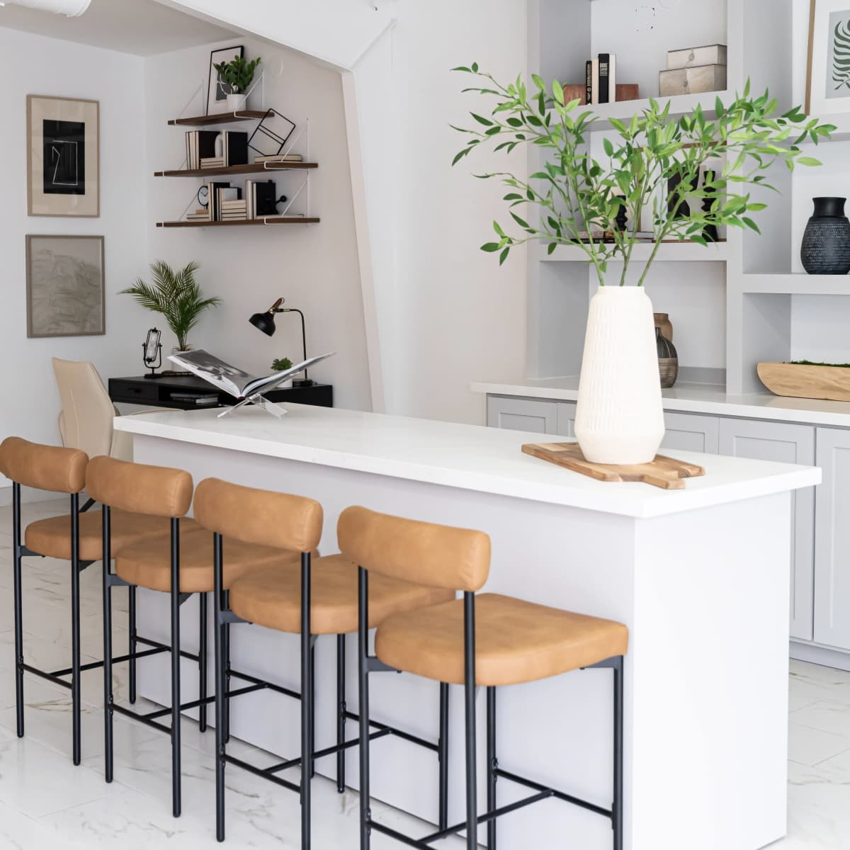 A modern kitchen features a white island with six tan, high-backed stools and a shelf holding decorative items in the background.