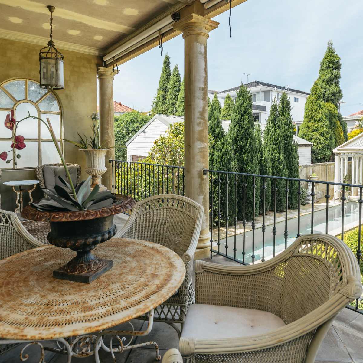 A covered balcony features a round table with chairs and a potted plant, overlooking a landscaped garden with a swimming pool and a white gazebo.