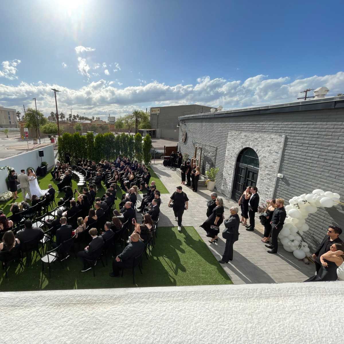 A wedding ceremony takes place outdoors with numerous seated guests facing a couple standing at the front, under a blue sky with scattered clouds.