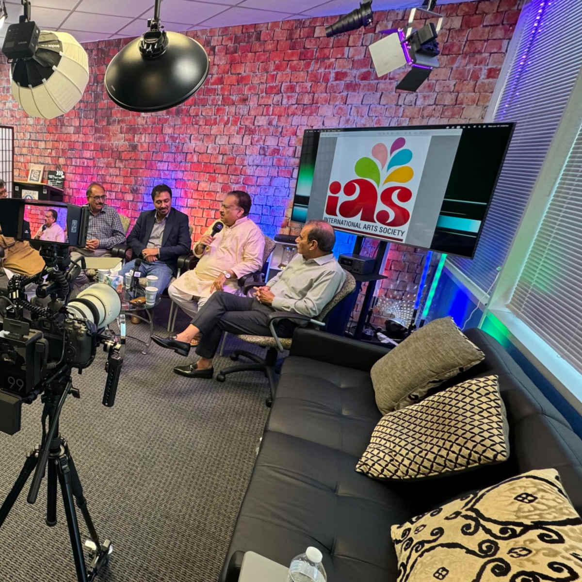 A group of five individuals is seated in a studio setting with multiple cameras and lights, discussing in front of a screen displaying a colorful logo.