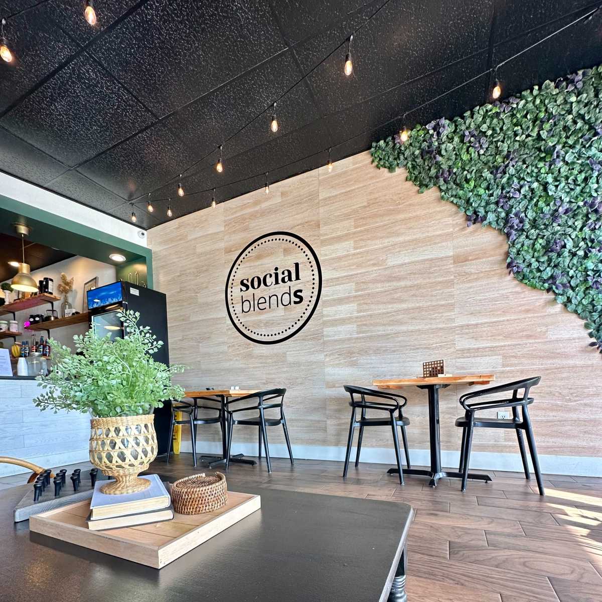 The interior of a café features tables and chairs near a wall adorned with greenery and a sign that reads "social blends."