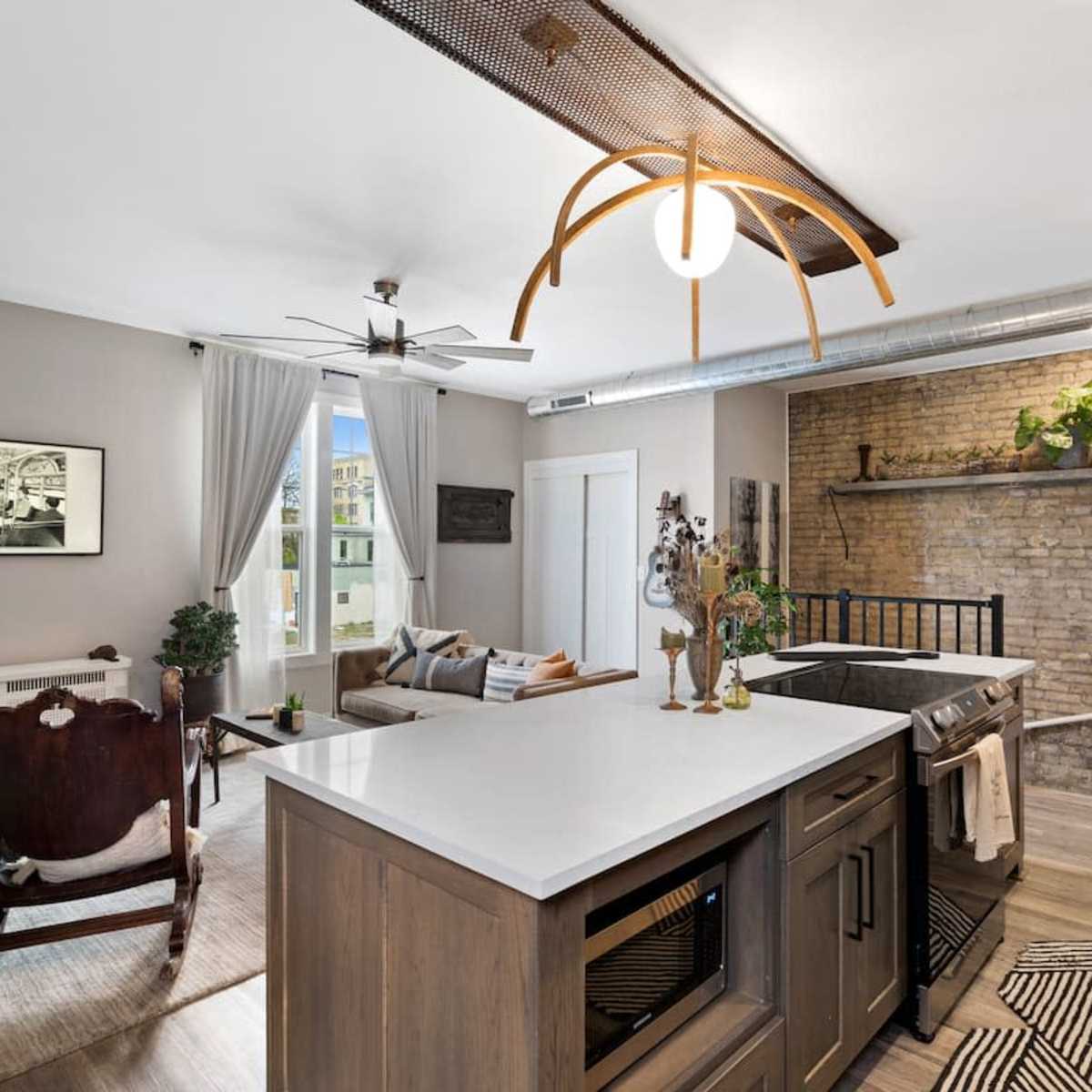A modern kitchen with contrasting wooden cabinetry, a central island, and a brick wall, adjacent to a cozy living area with a vintage chair and stylish decor.