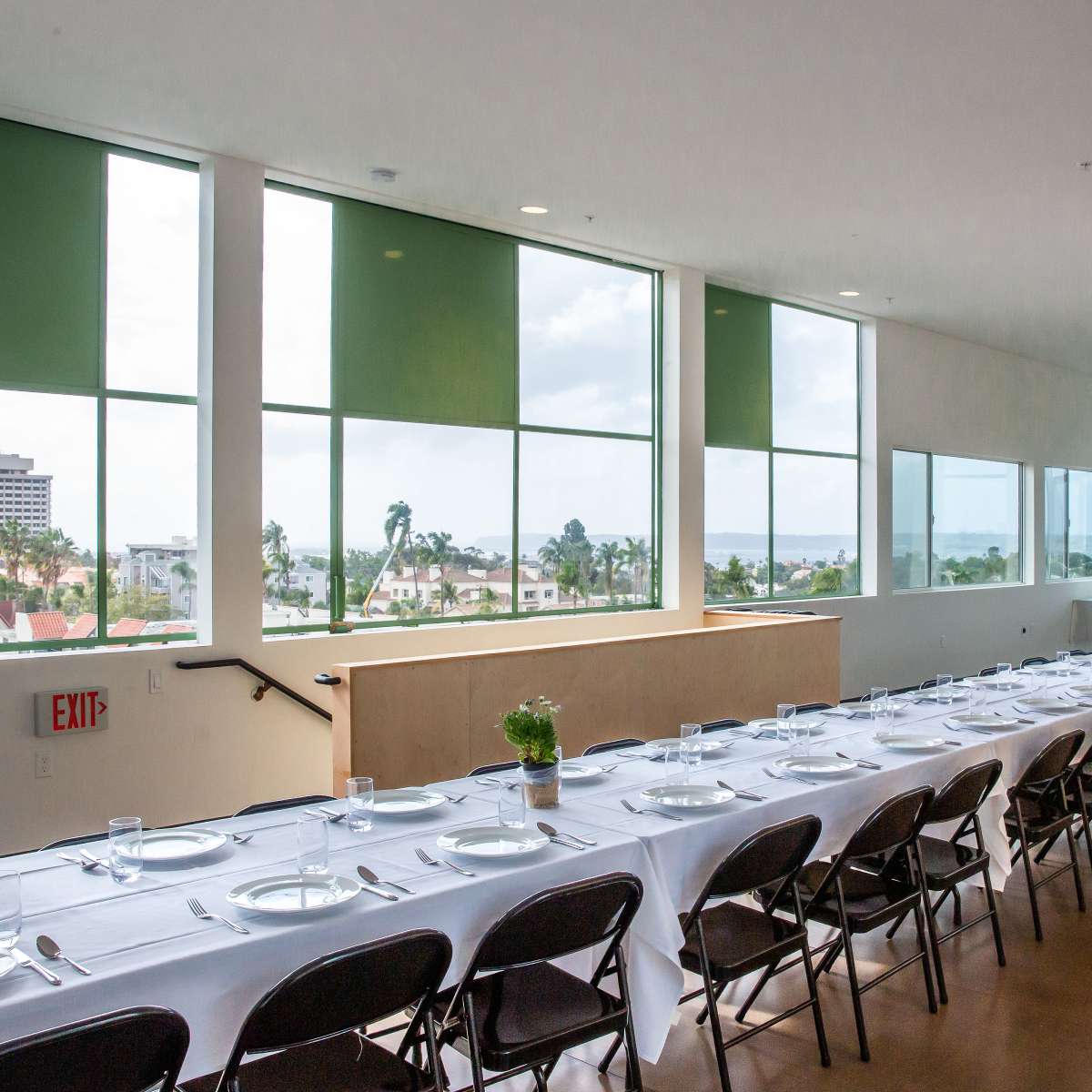 A long, elegantly set dining table with white tablecloths is arranged beside large windows overlooking a cityscape and the ocean in the background.