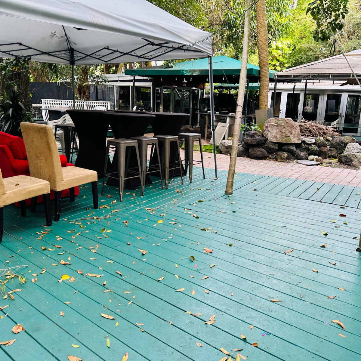 A wooden deck with scattered leaves features tables and chairs set up under canopies in a landscaped outdoor area.