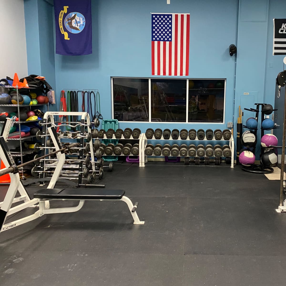 A gym interior with weightlifting equipment, exercise balls, and free weights lined up against the wall, adorned with American and military flags.