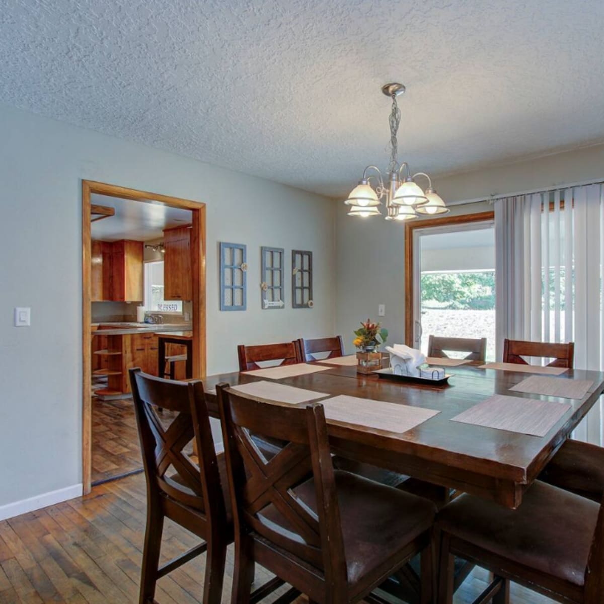 A spacious dining area features a large wooden table surrounded by eight chairs, with a hanging light fixture above and a view of the kitchen through an open doorway.