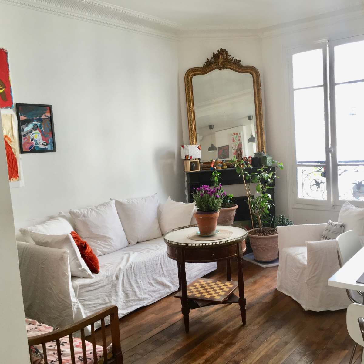 A bright living room features a white sofa and armchair, a circular table with a potted plant, and large windows with a view of a balcony.