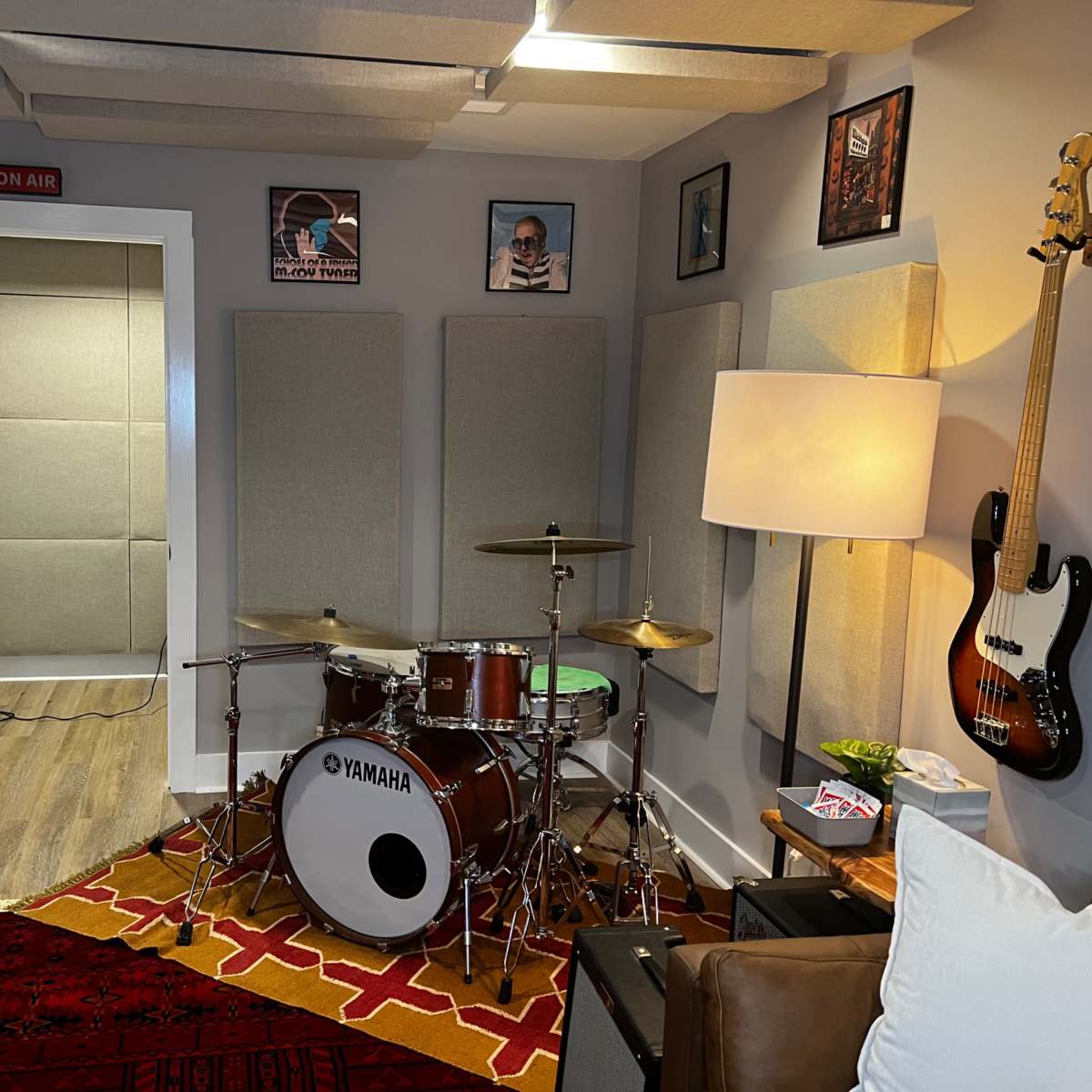 A music rehearsal room featuring a drum set, a bass guitar, and an adjacent soundproofed room with a door labeled "ON AIR."