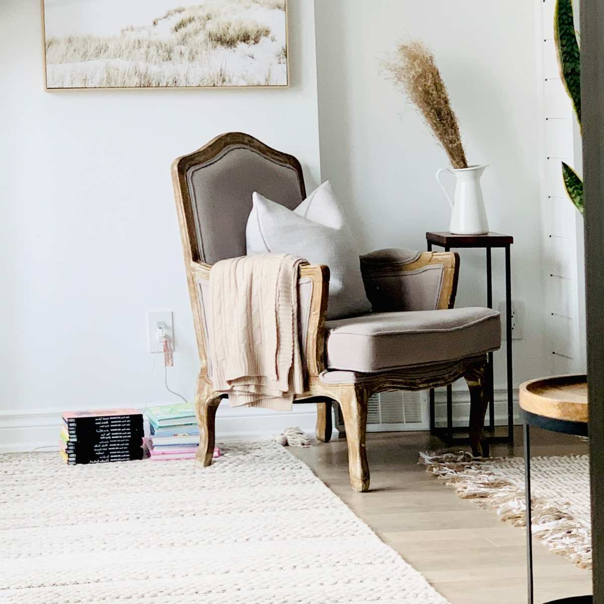 A vintage armchair with a throw blanket sits beside a small side table and a decorative vase in a bright, minimalistic room.