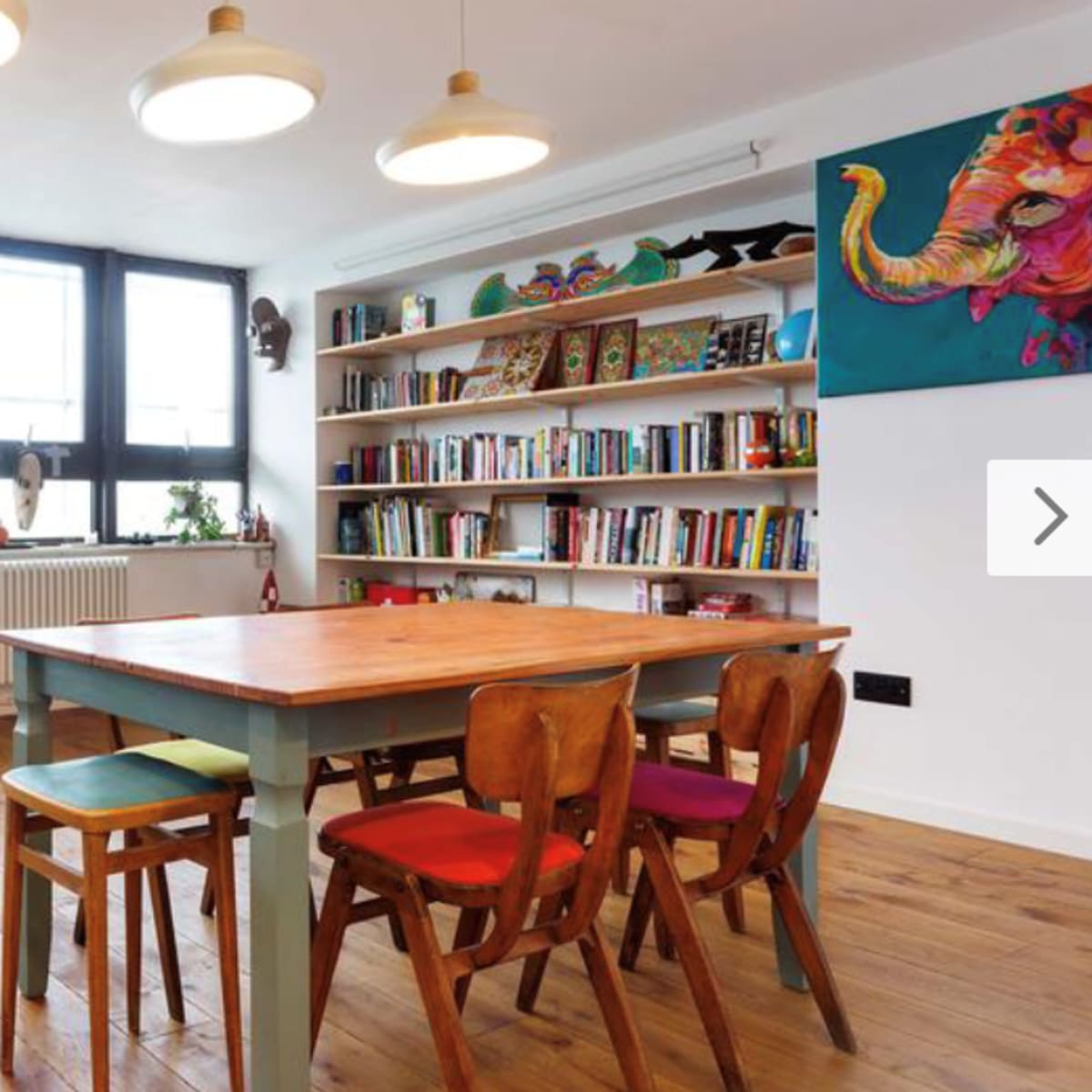 A bright, modern living room with a wooden dining table surrounded by colorful chairs, a large bookshelf filled with books, and an elephant painting on the wall.