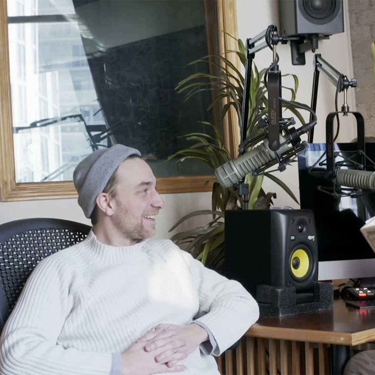 Two men sit in a studio setting, one reading from a book while the other listens, surrounded by audio equipment and a plant.