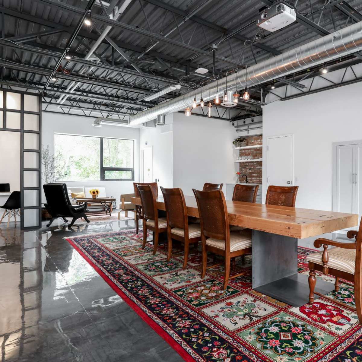 A modern interior space featuring a long wooden dining table surrounded by eight chairs, placed on a colorful area rug, with large windows and metal ductwork visible in the ceiling.