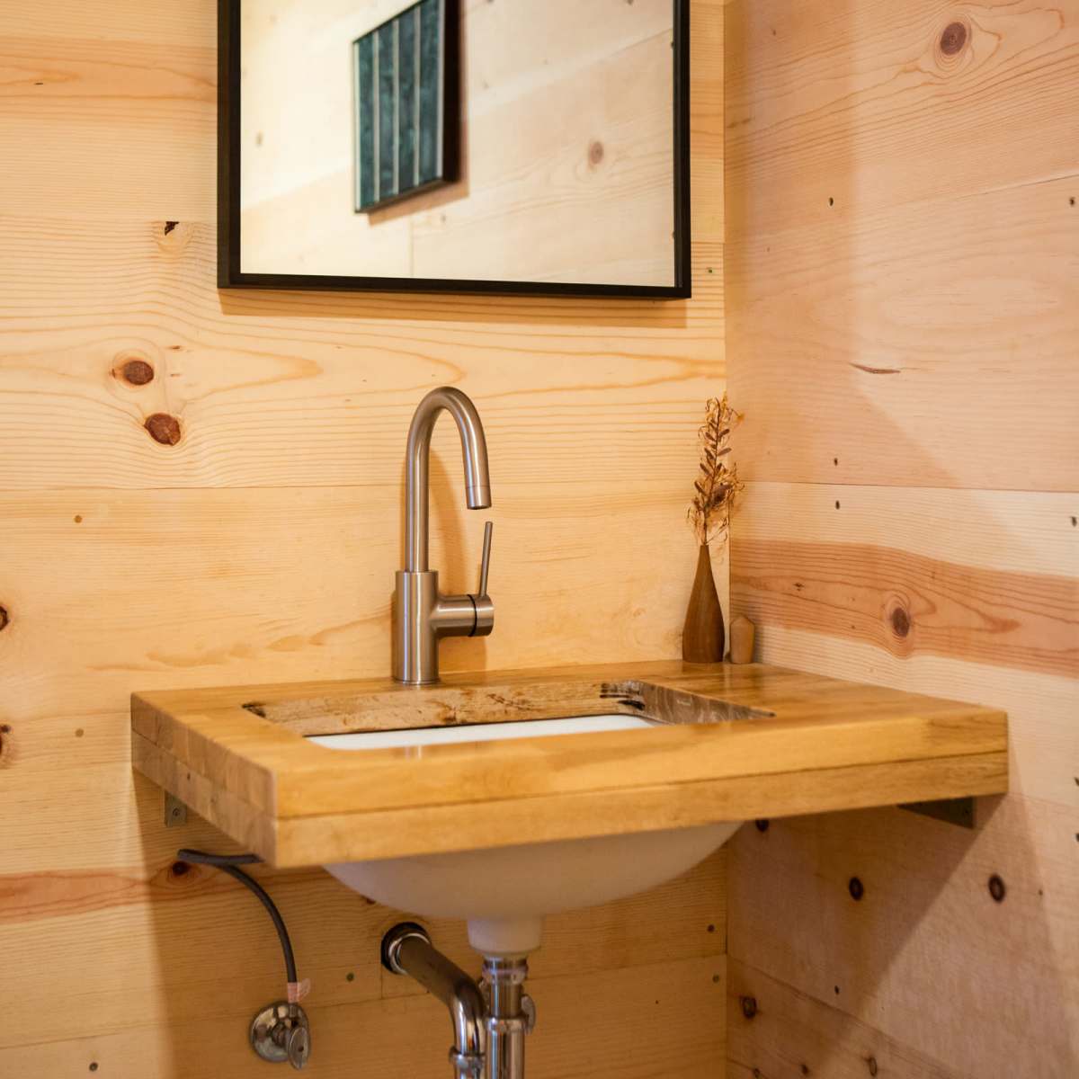 A wooden sink with a minimalist faucet is mounted on a wall with light-colored wood paneling, reflecting a simple framed artwork in the mirror above it.