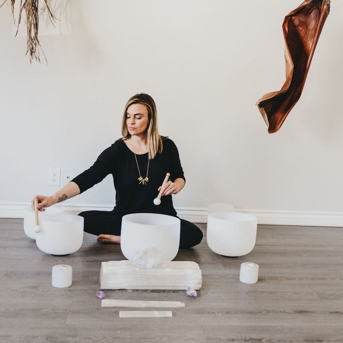 A woman sits on the floor, playing crystal singing bowls, with decorated wall elements made of dried leaves above her.