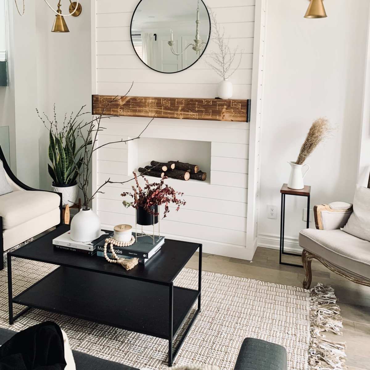 A modern living room with a white shiplap wall featuring a round mirror above a wooden mantel, a simple fireplace, and two seating areas, all decorated with plants and minimalistic decor.