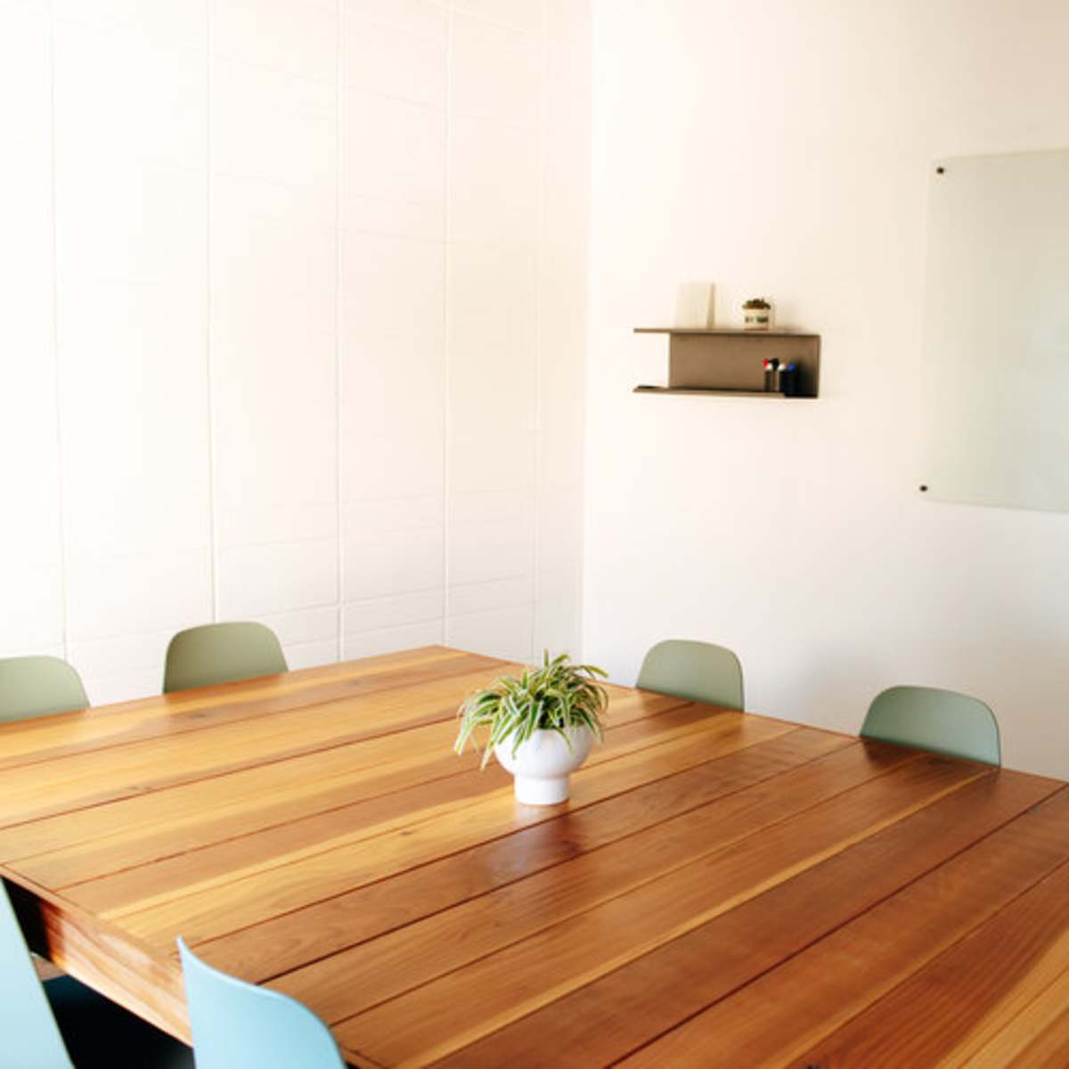 A wooden conference table surrounded by light green chairs sits in a well-lit room with a whiteboard mounted on the wall.