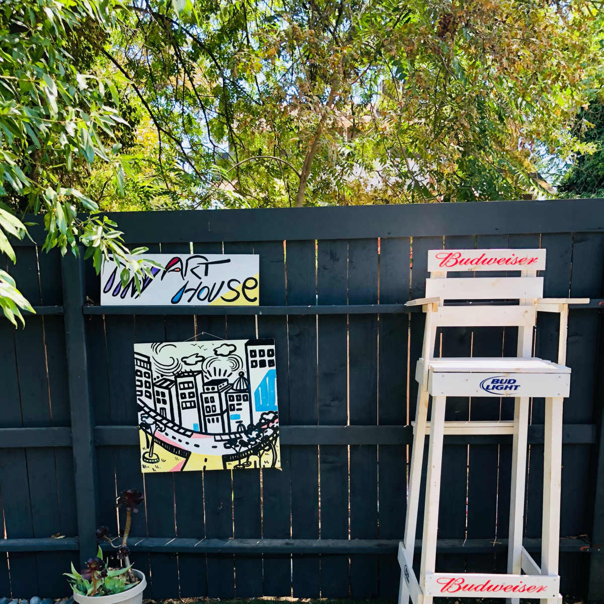 A wooden fence displays a colorful art piece and a tall white stool beside a small potted plant on a grassy area.