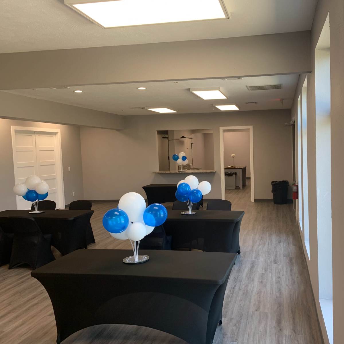 A spacious event room with black tables decorated with blue and white balloon centerpieces, large windows, and a serving area in the background.