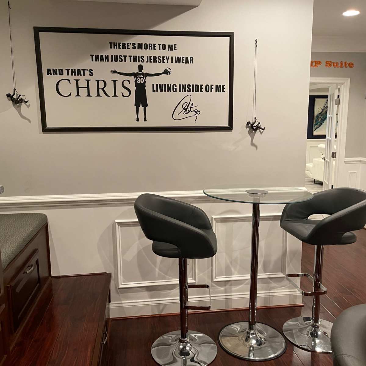 A small seating area with a glass table and two black bar stools, surrounded by white walls featuring a framed wall art depicting a jersey and the name "CHRIS."