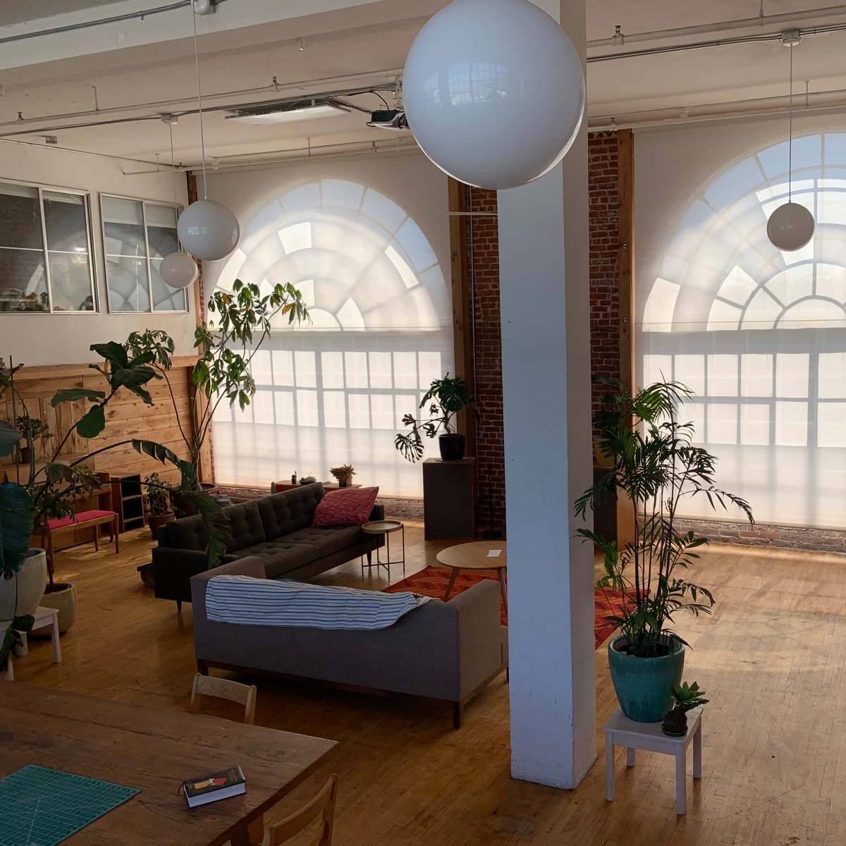 The room features large arched windows, a cozy seating area with a sofa and coffee table, and several indoor plants on wooden floors.