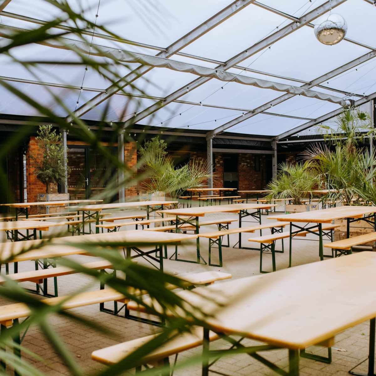 An outdoor event space features numerous wooden tables arranged under a transparent roof, surrounded by potted plants.