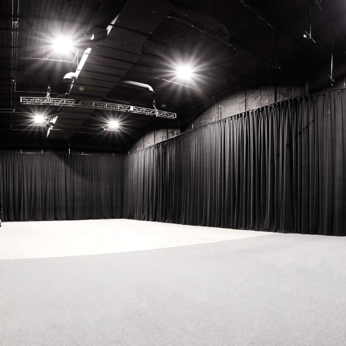 A spacious studio with black curtains lining the walls and bright overhead lights illuminating the gray floor.