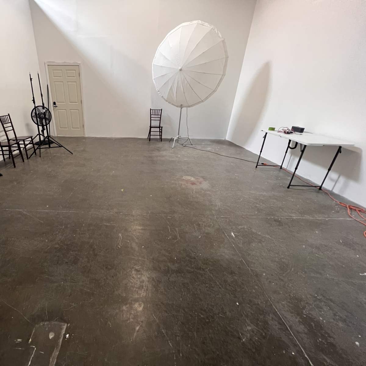 A spacious, empty studio with a folding chair, a large lighting umbrella, a table, and a few other chairs against the walls.