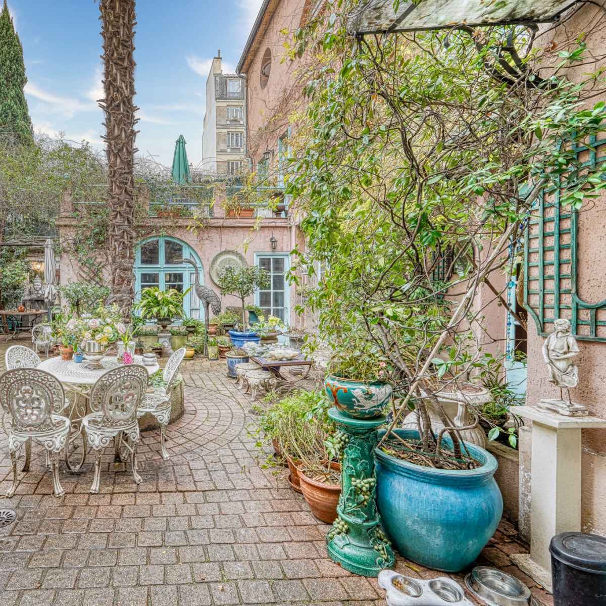 A lush courtyard garden with various potted plants, stone pathways, and a table set with chairs under a palm tree.