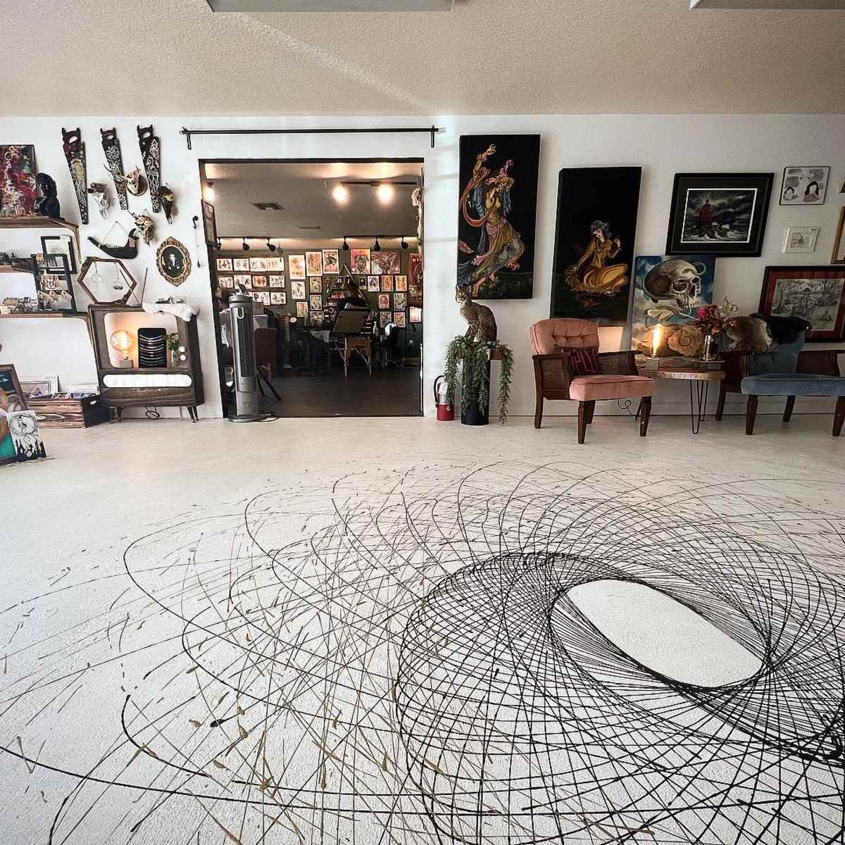 An art studio with a spiraling black pattern on the white floor, surrounded by various framed artworks and decorative items on the walls.