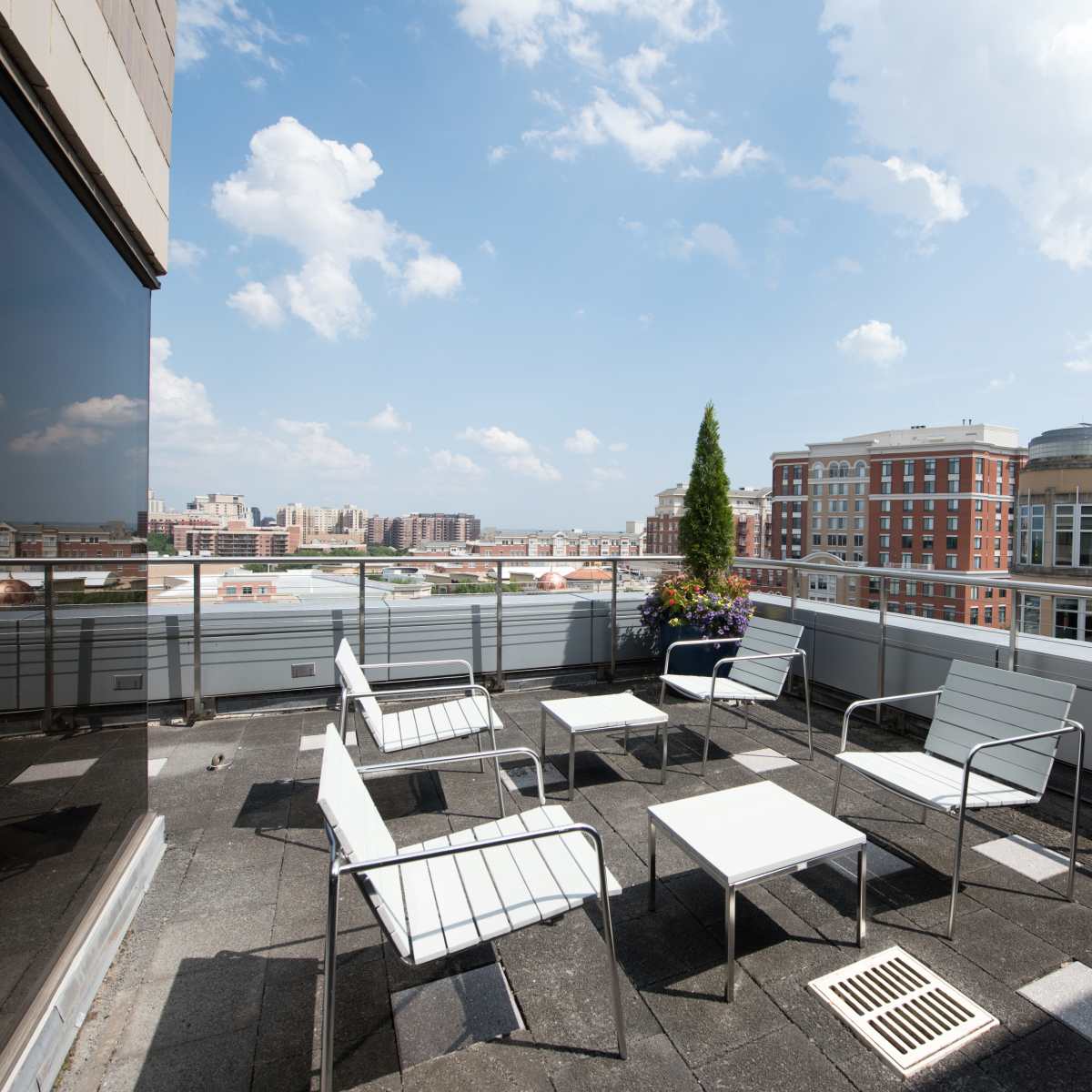 A rooftop terrace features several white chairs and tables, overlooking a city skyline under a partly cloudy sky.