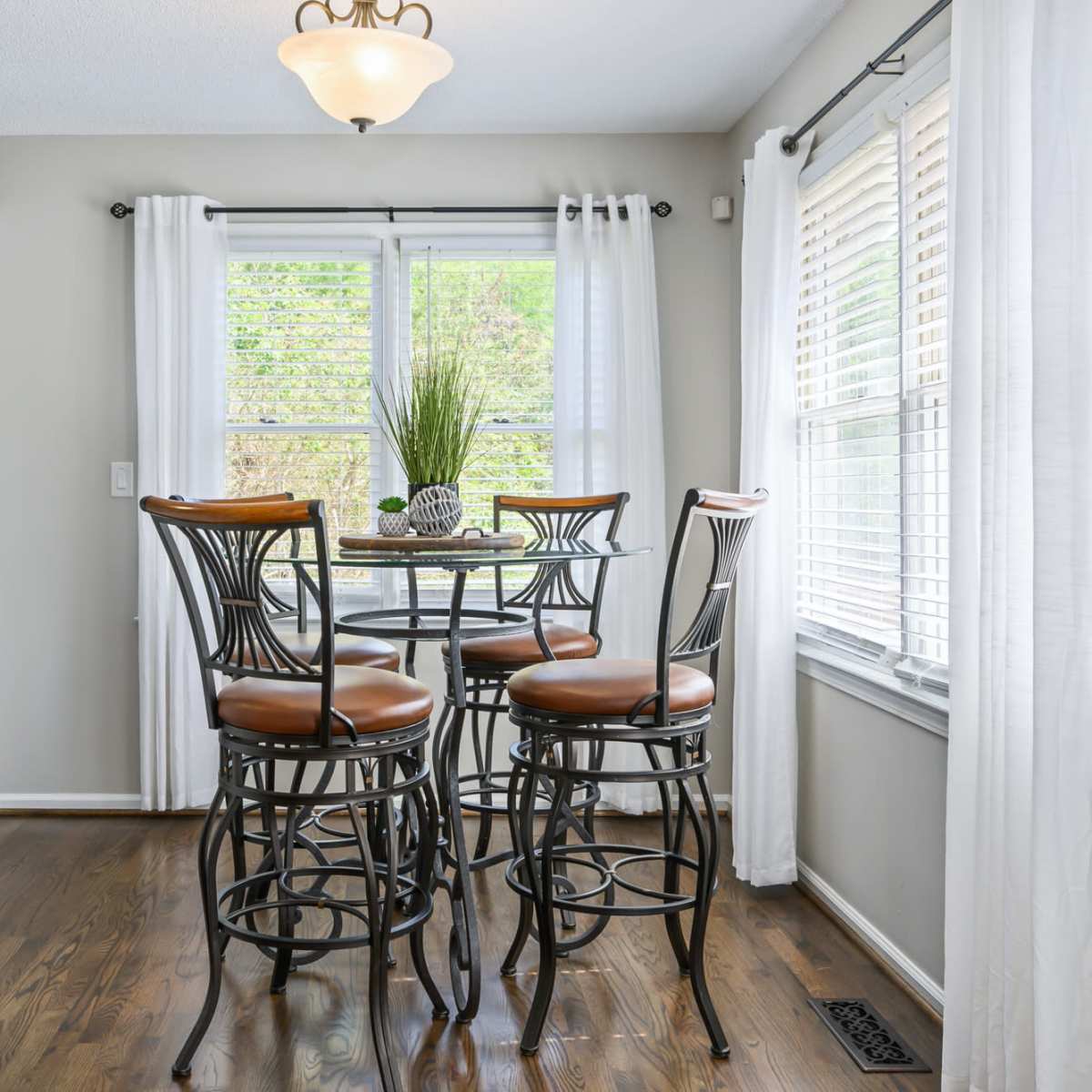 A breakfast nook features a round glass table surrounded by four metal bar stools, with large windows letting in natural light and potted plants as decor.