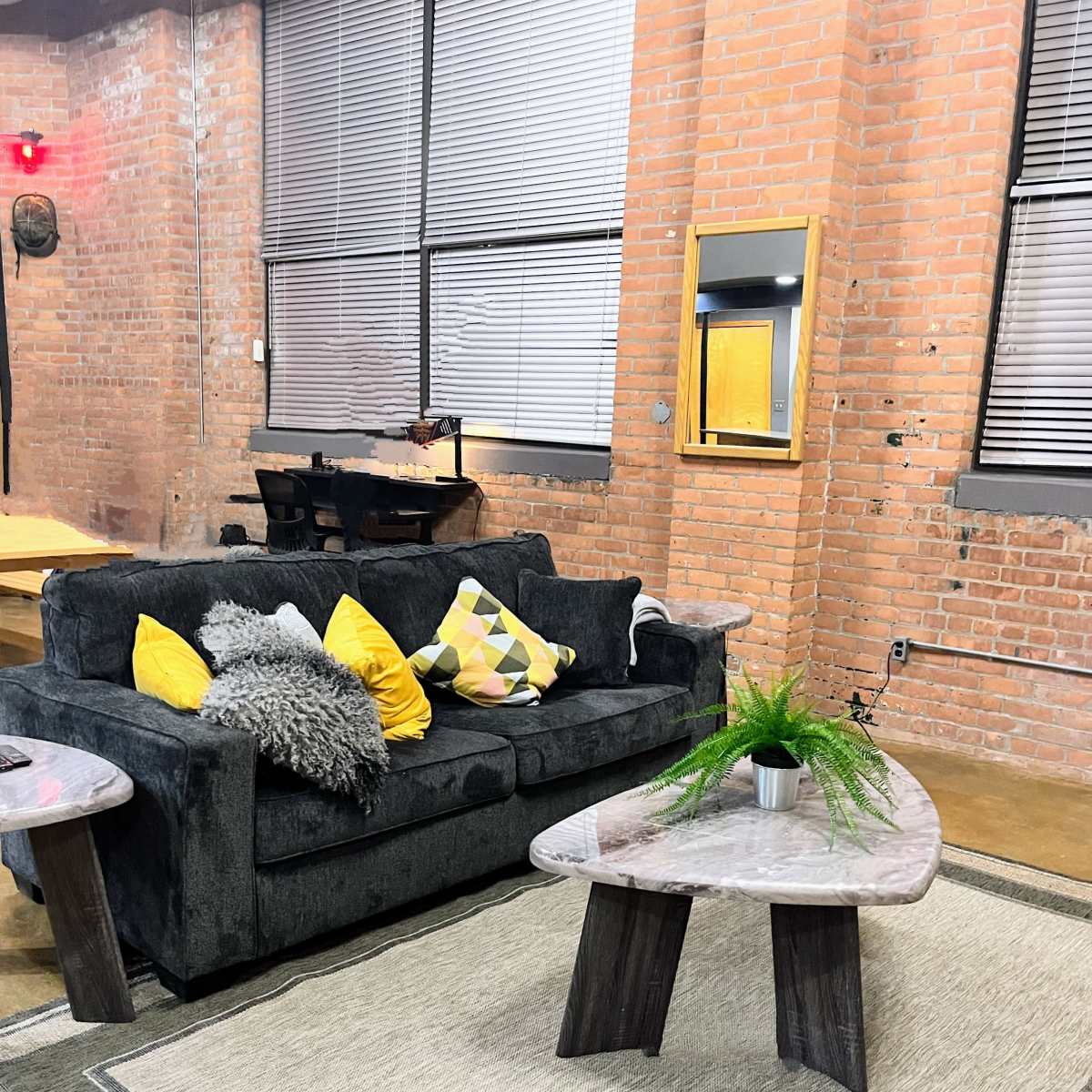 A living room with a dark sofa adorned with yellow and patterned cushions, a round coffee table, and a small plant on a textured area rug, all set against exposed brick walls.