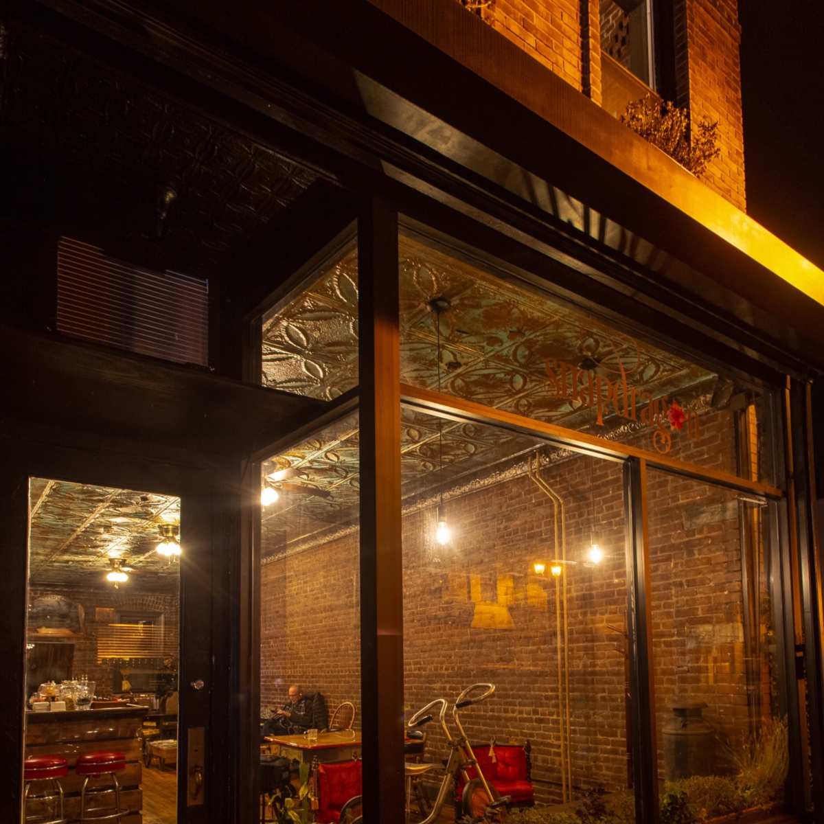 The exterior of a restaurant with large windows, revealing an interior with brick walls and red seating, illuminated by pendant lights at night.