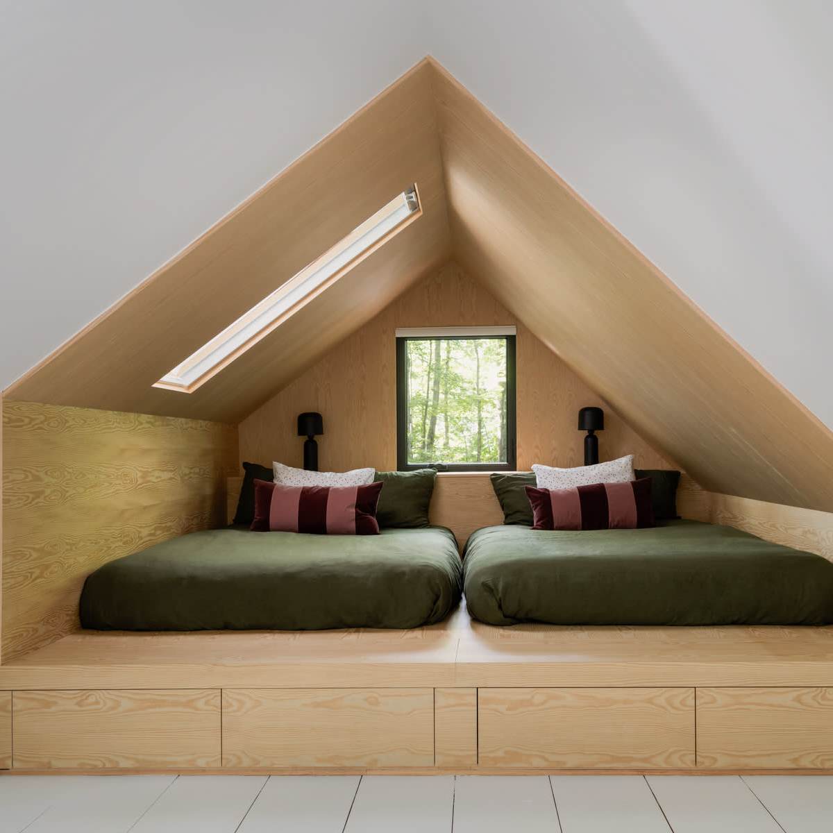 An attic bedroom with two green beds placed on a wooden platform under a sloped ceiling, featuring a window that overlooks trees.