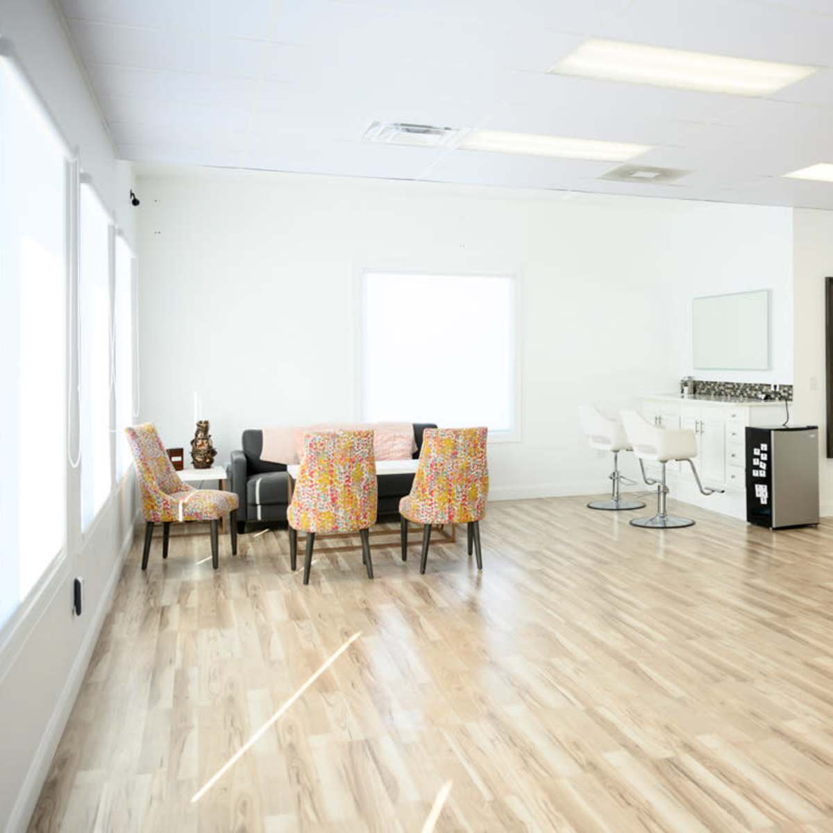 A spacious, well-lit room with a seating area featuring two patterned chairs, a bar counter with stools, a mirror, and a door leading to another space.