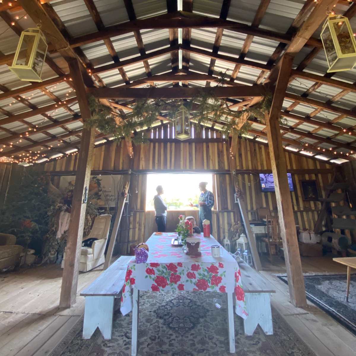 The interior of a rustic barn features a table with a floral tablecloth and two people standing by a window, surrounded by wooden beams and decorative lights.