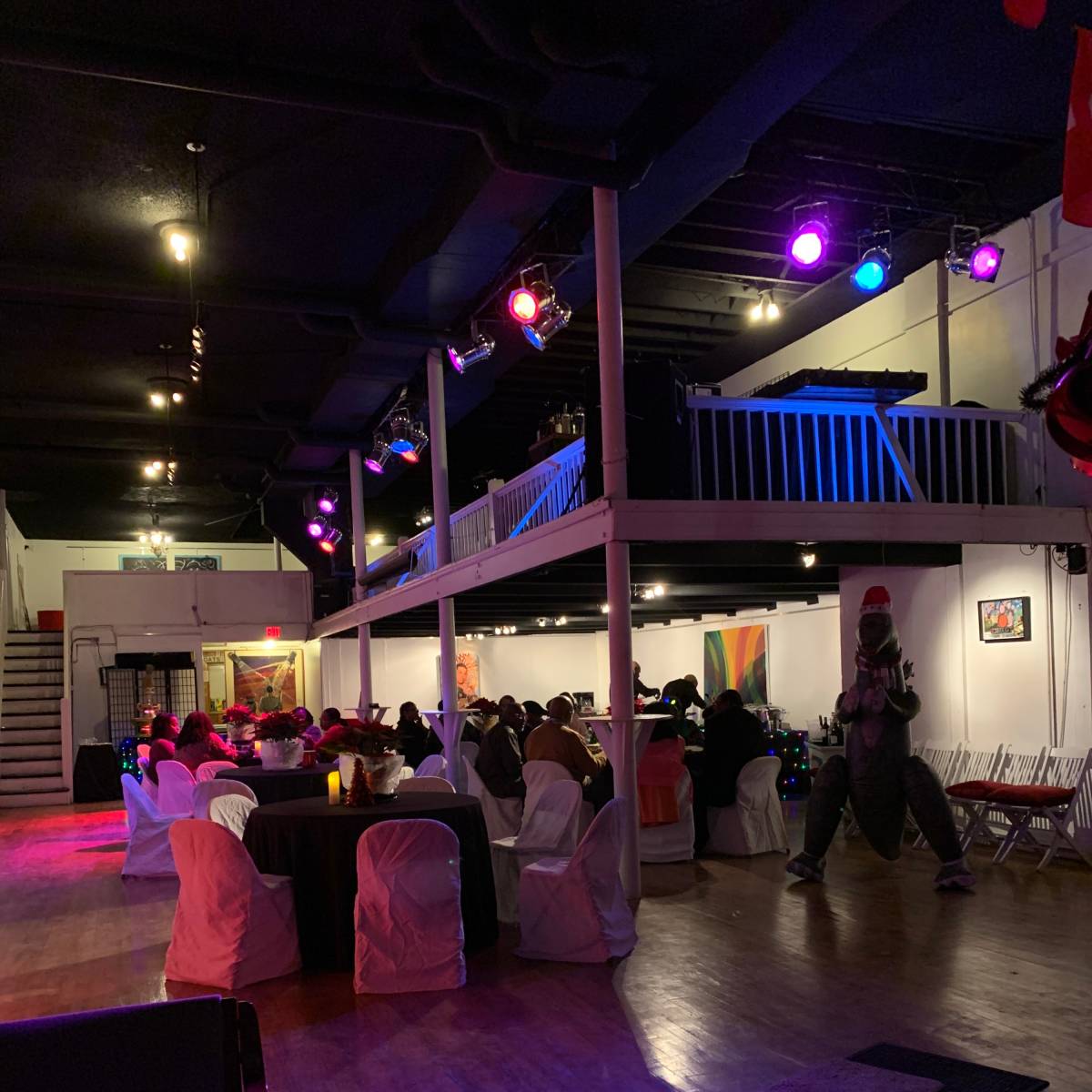 A spacious event venue with guests seated at tables, illuminated by colorful lights, and a person in a costume standing to the side.