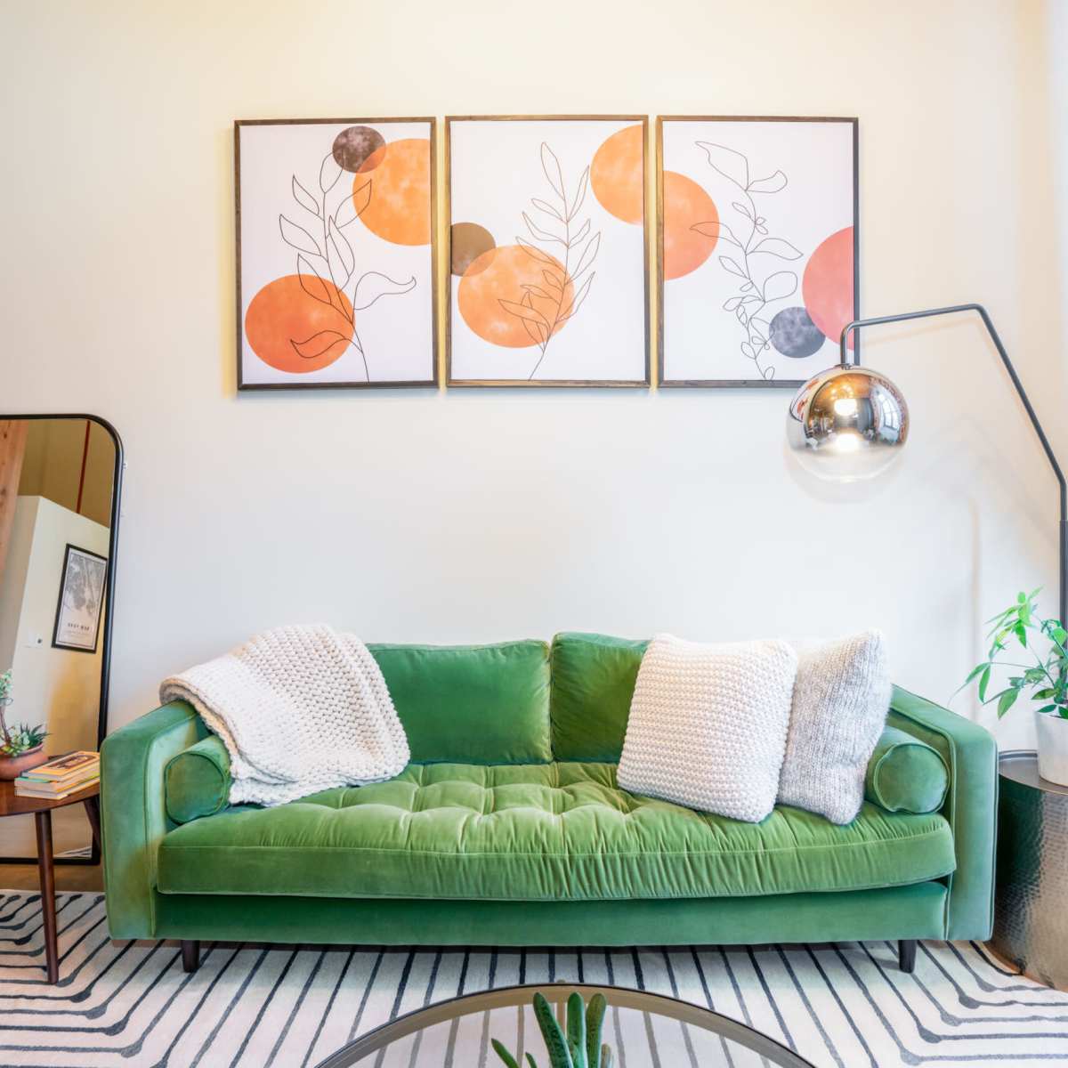 A green velvet sofa with two white pillows, set against a wall featuring three framed abstract artworks, next to a round coffee table and a large mirror.