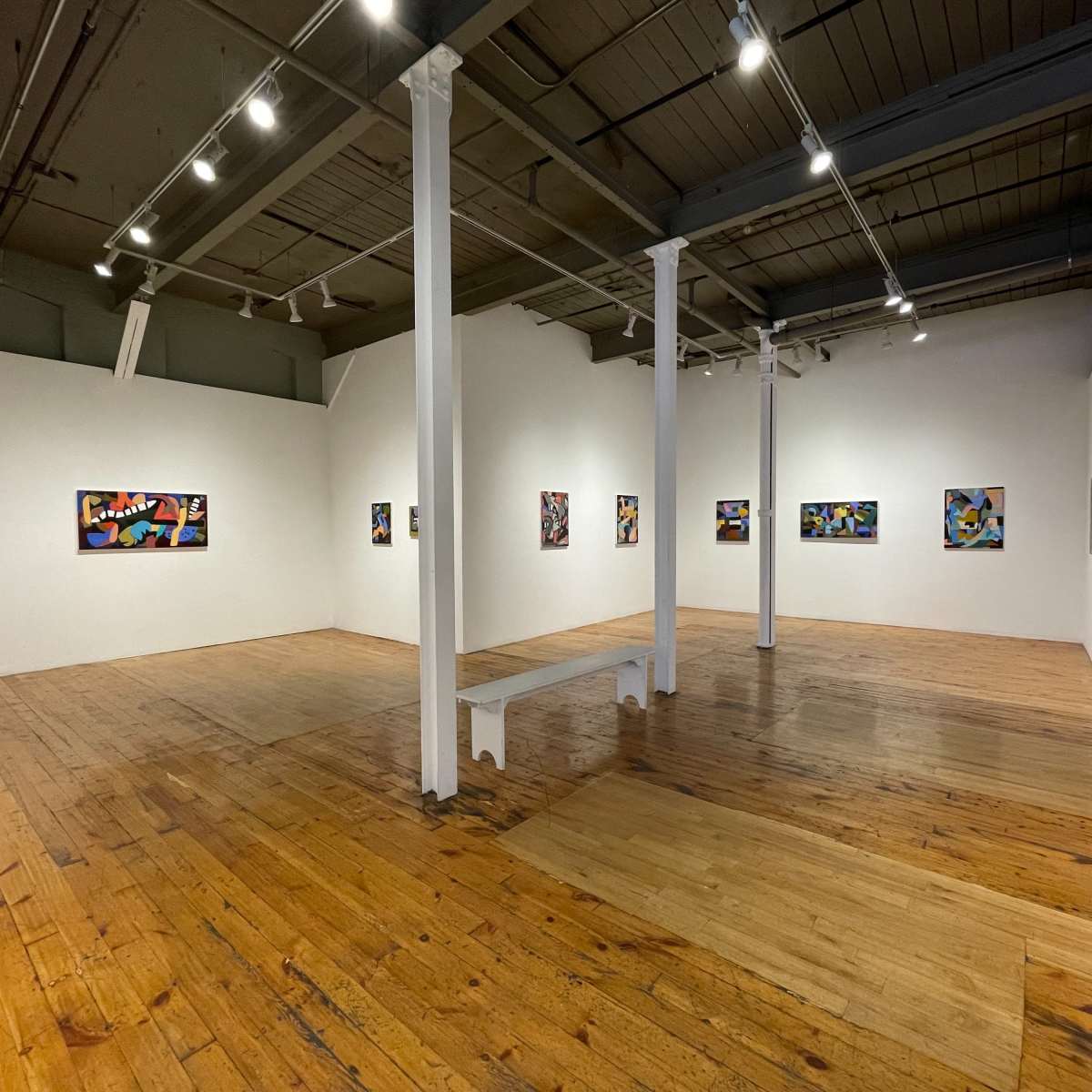 A spacious art gallery with white walls, wooden flooring, and several colorful paintings displayed along the walls.