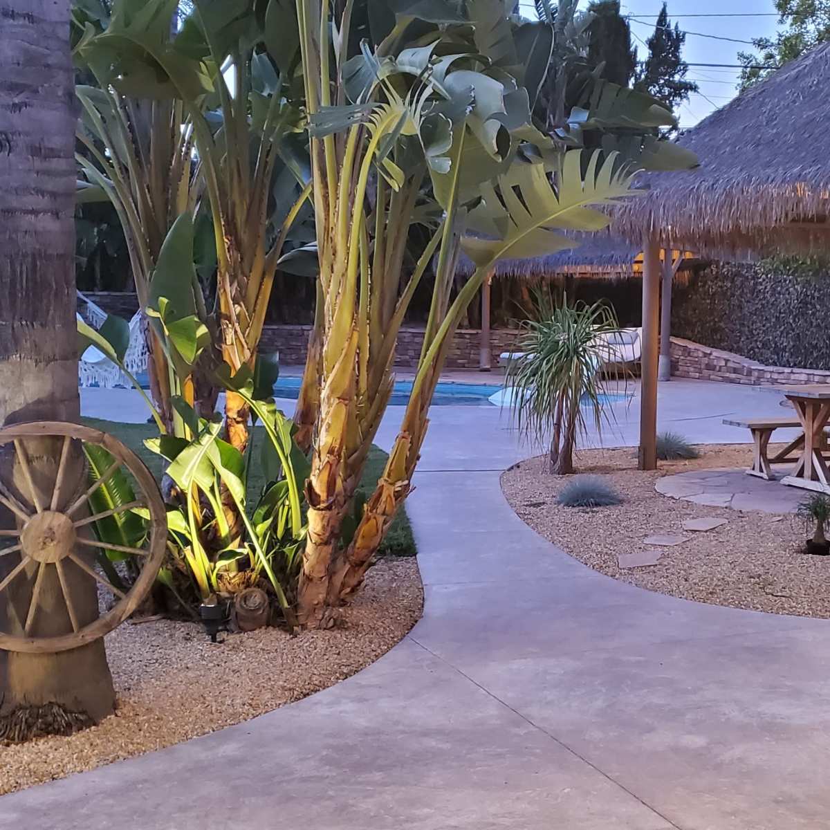 A backyard with a swimming pool, a thatched-roof gazebo with picnic seating, and tropical plants surrounding a stone pathway.
