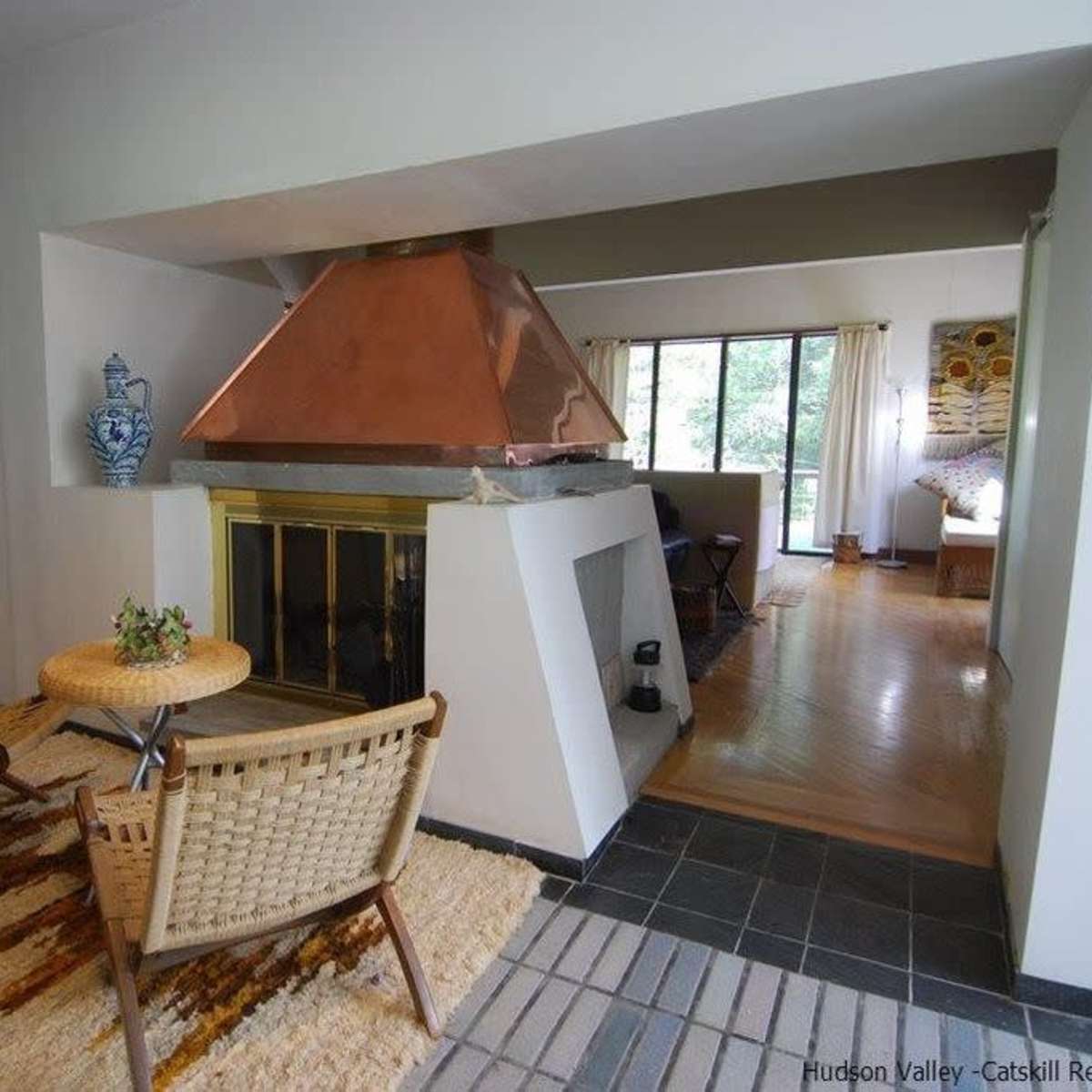 A modern living space featuring a copper chimney above a fireplace, surrounded by two woven chairs and a small table, with large windows allowing natural light.