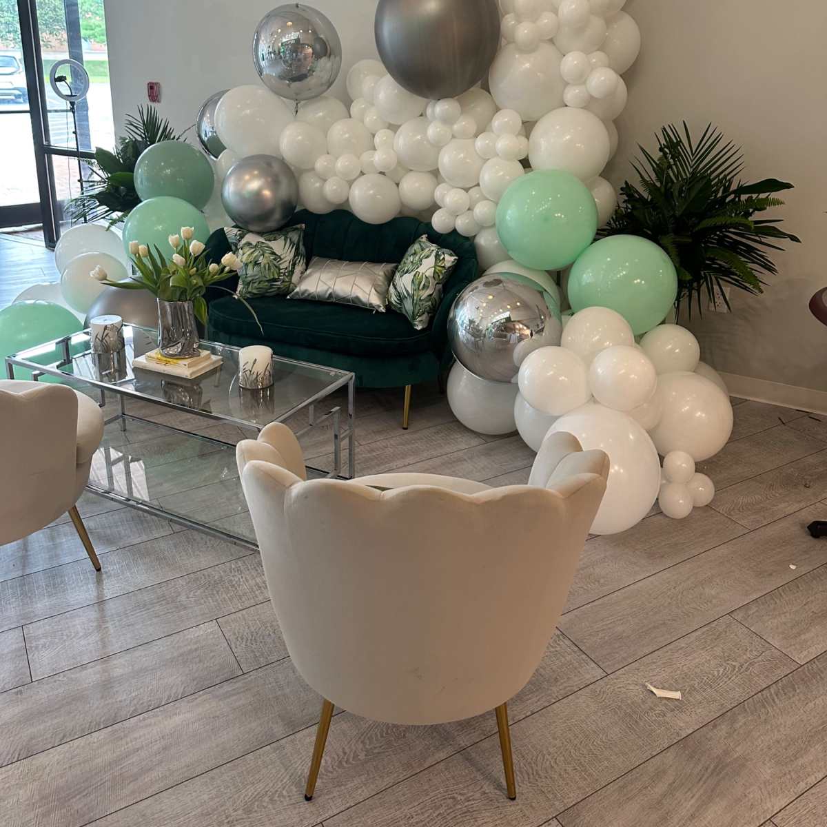 A cozy seating area with a green couch surrounded by a decorative arrangement of white and mint balloons, silver spheres, and potted plants.